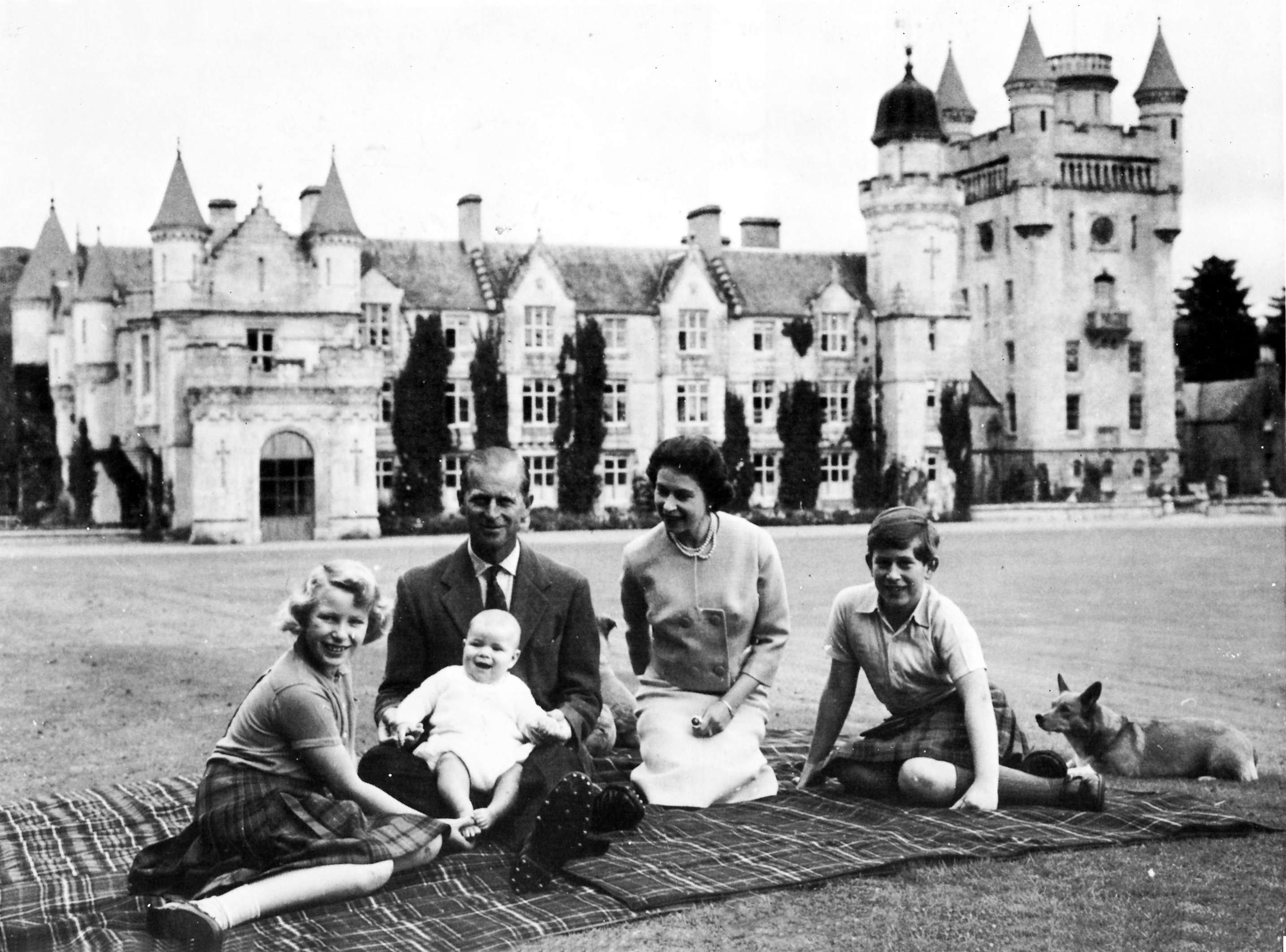 Queen Elizabeth and Prince Philip sitting on a blanket outside Balmoral Castle with baby Prince Andrew and young Prince Charles and Princess Anne