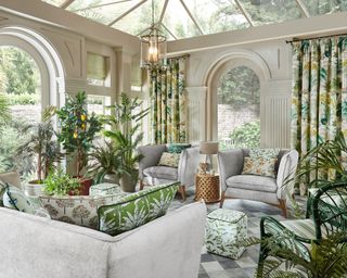 Orangery with columned, curved window by ILIV – a room full of plants. grey sofas and leafy accessories such as footstools.