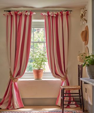 A window with two cream and dark pink striped curtain panels draped either side and a terracotta plant pot on the ledge, three hats on the righthand wall, and a black wooden chair underneath these