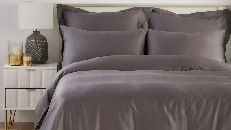 A luxurious set of dark charcoal grey bamboo bedding is neatly arranged on a bed next to a white modern nightstand with a lamp and candles.