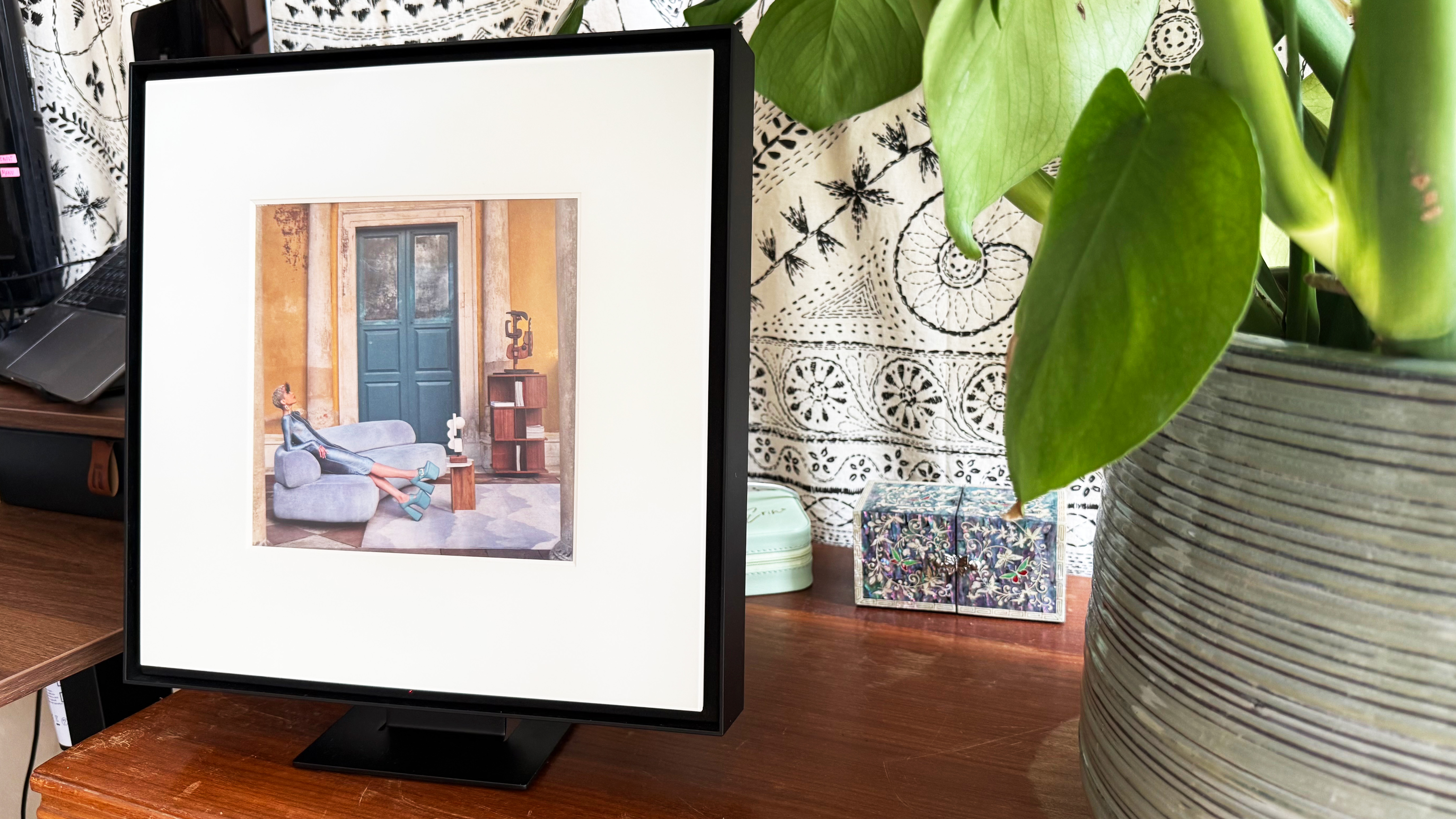 the samsung music frame close up with a green plant beside it and a white and black tapestry behind