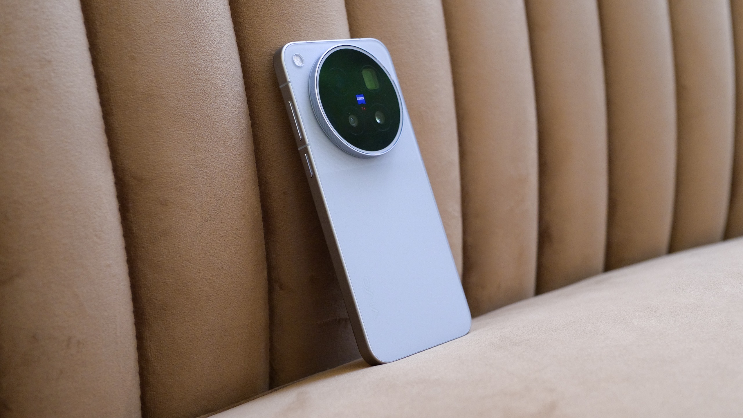 Vivo X500 Pro Max leaning against a tan upholstered bench, showing its slim silver body and circular rear camera module.