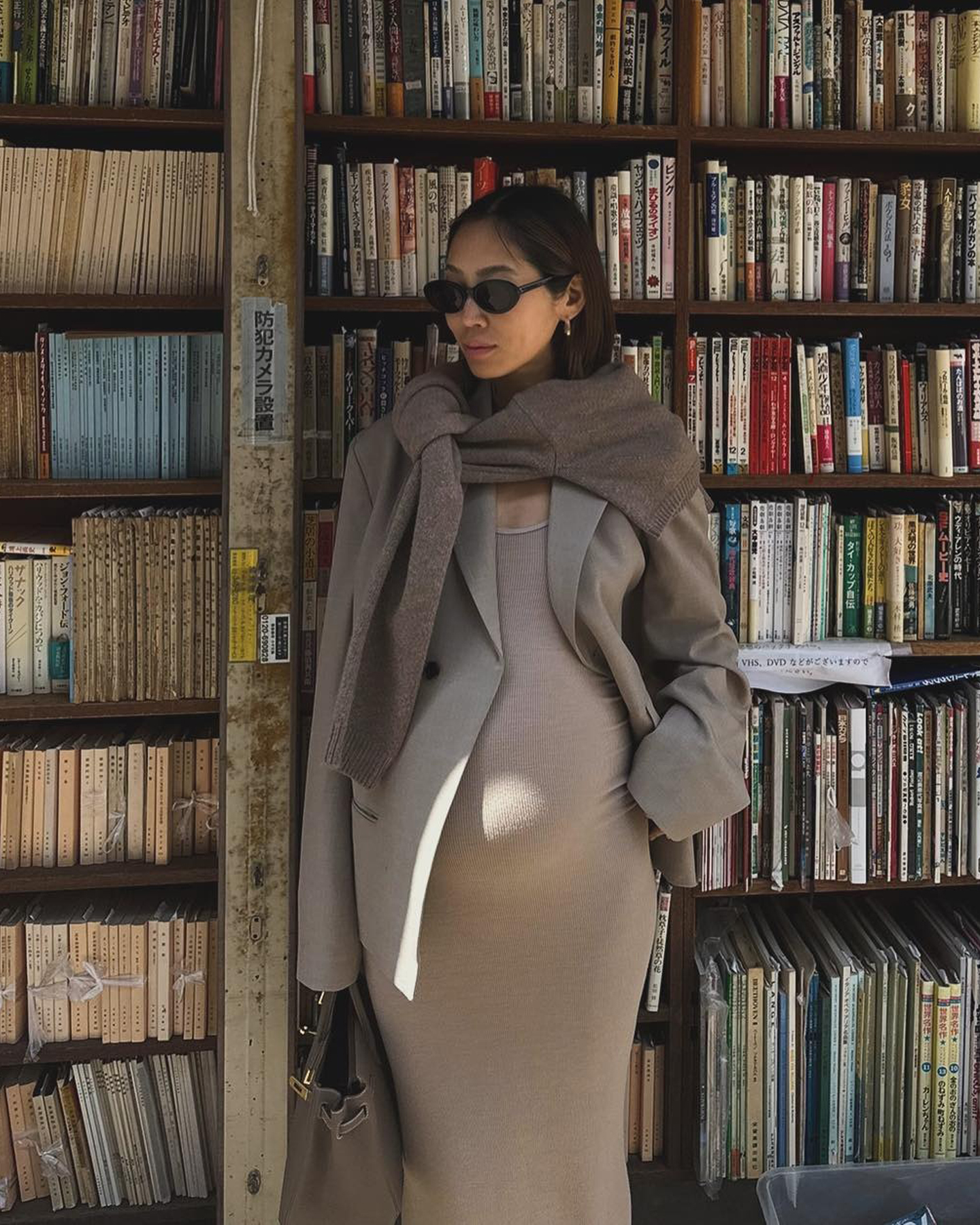 A pregnant Aimee Song poses in front of a full bookshelf wearing a chic maternity outfit with black oval sunglasses, a brown sweater tied around her shoulders, a neutral blazer, tan knit dress, and a designer bag