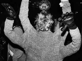 Black and white photo of someone who has mask with two stitched faces, four arms but only one eyeball