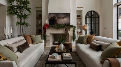 bright living room with two white sofas facing a stone fireplace decorated with evergreen garland, red ribbons, and a painting of Santa and his sleigh