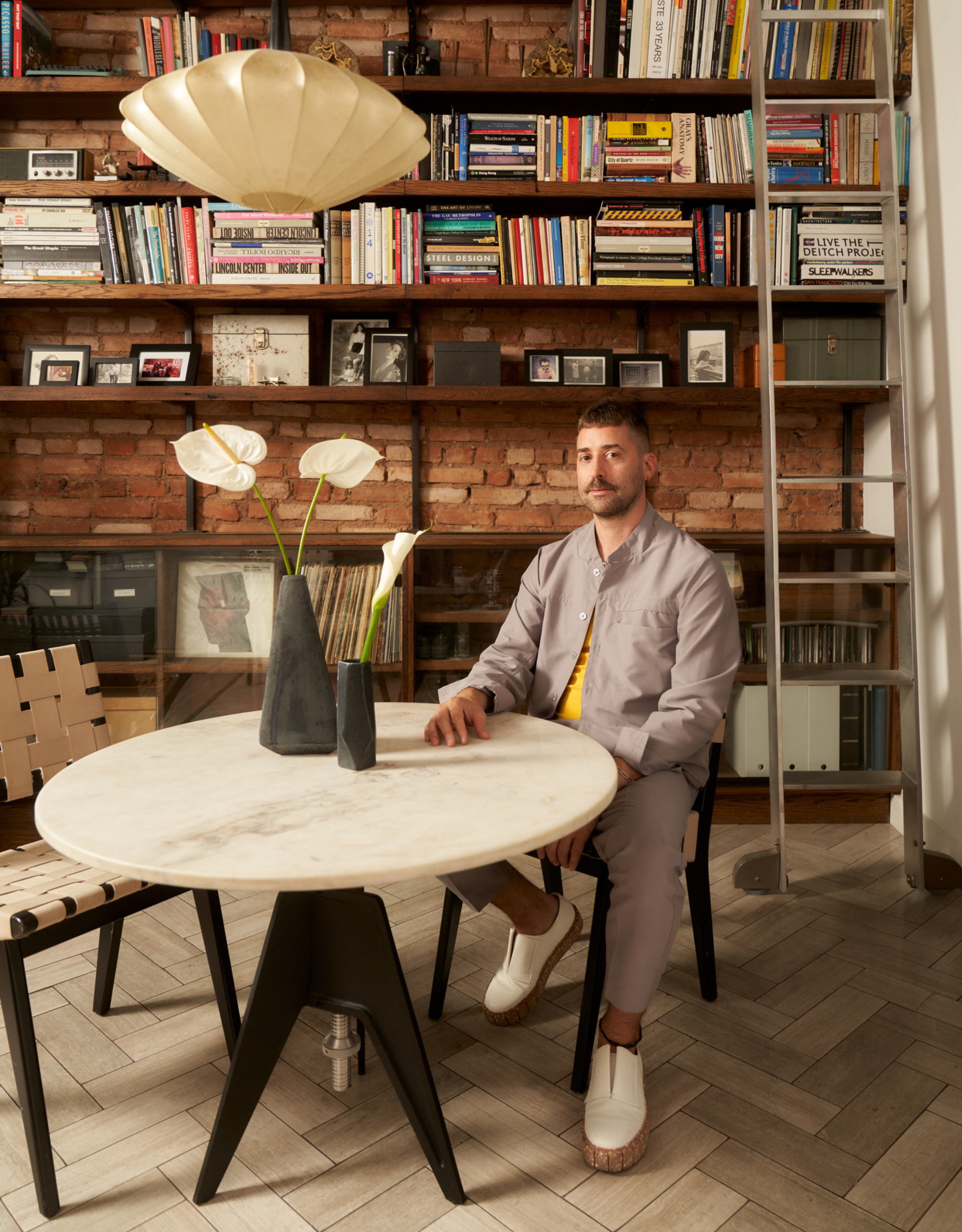 An NYC loft with a bookshelf against a brick wall, a dining table with a black chair with leather detailing, and a pendant light. A man in a gray co-ord with white shoes is seated at the table 