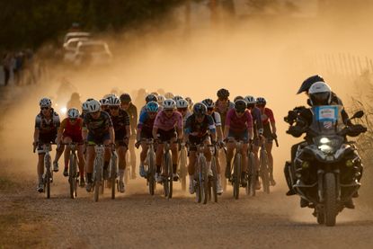 A peloton of men on gravel bikes ride on a red, dusty road behind a lead motorcycle.