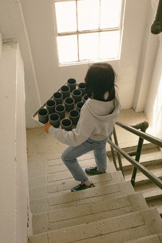 Image of a woman in jeans and a gray hoodie walking down an all-white staircase. The woman is holding a silver tray full of dark gray ceramic mugs.