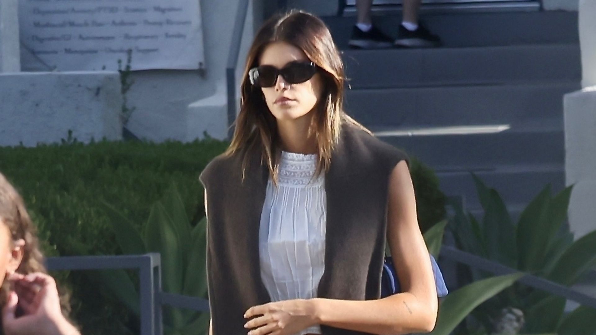 Kaia Gerber walks down the street wearing navy trousers and burgundy ballet flats with a white short sleeve blouse and a brown knit over her shoulders.