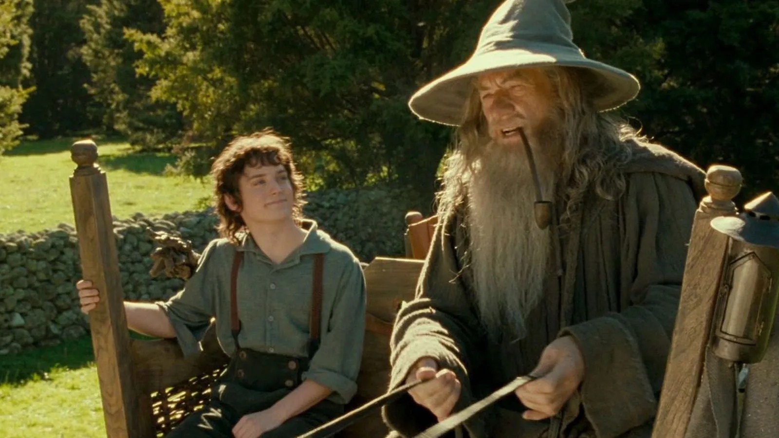 Still image from Lord of the Rings. Bilbo and Gandalf sit on  a cart on a sunny day in Hobbiton.