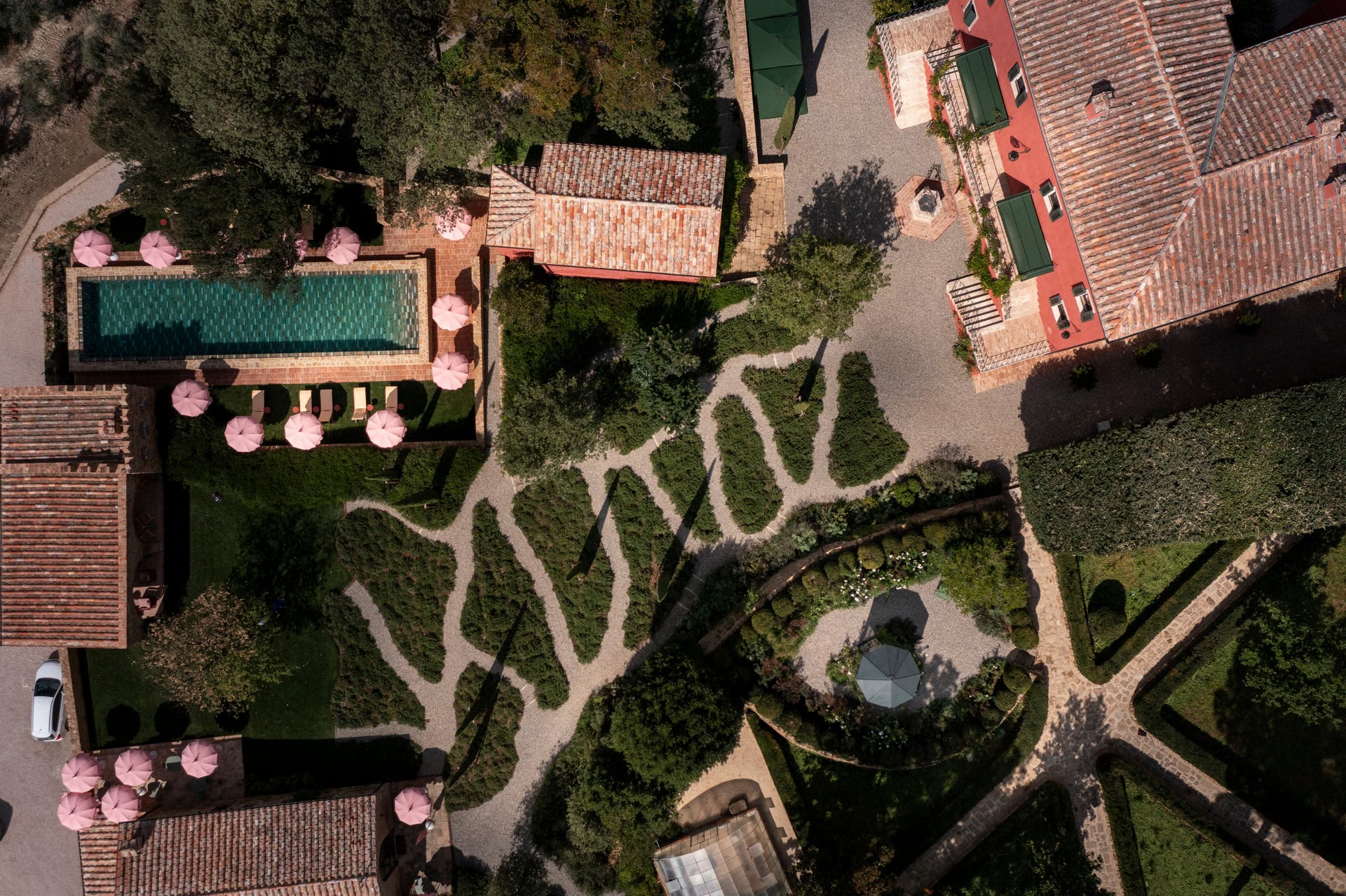 An aerial view of a hotel's gardens, dotted in pink parasols and lush patches of green, and surrounded by red tile ceilings.