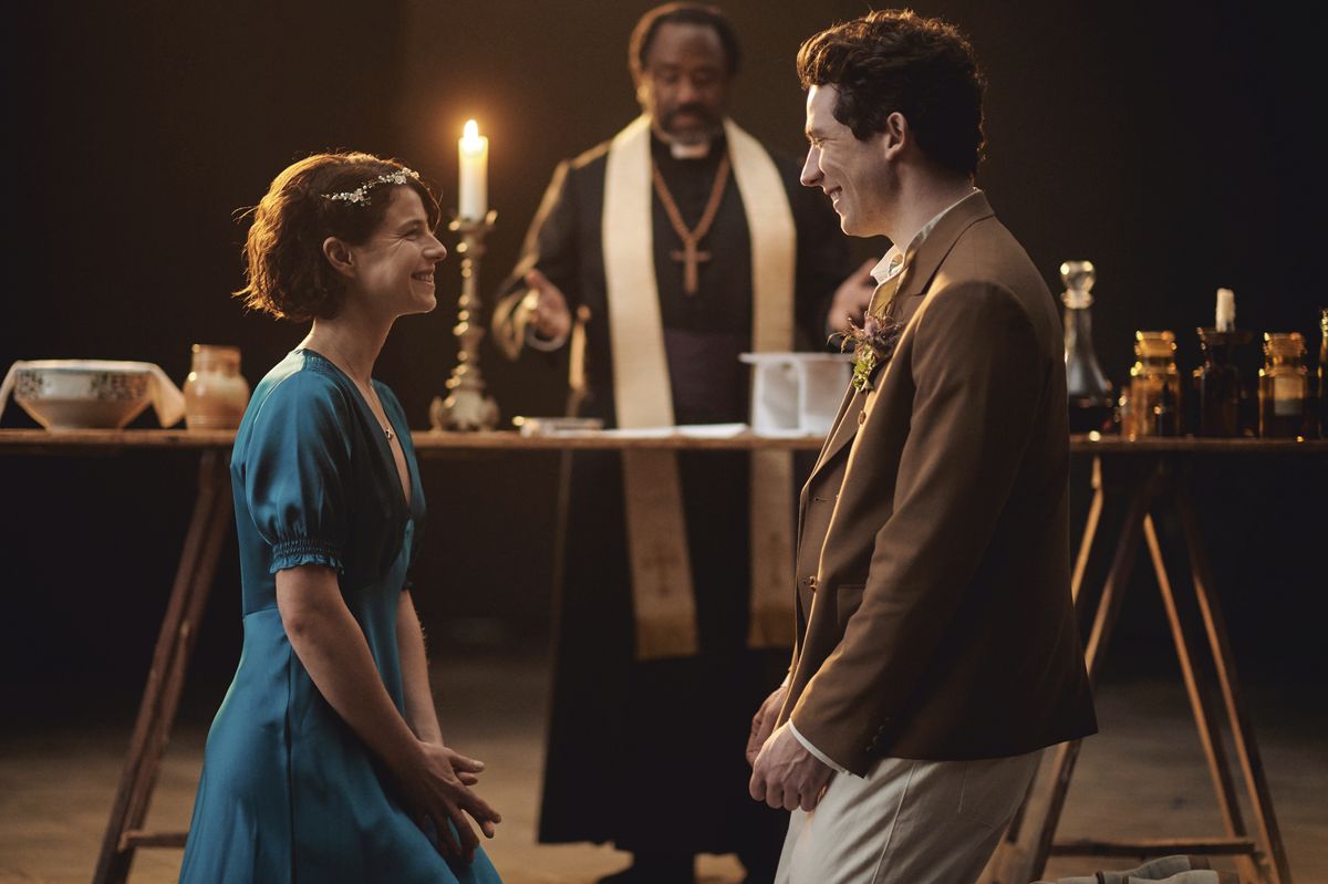 Juliet (Jessie Buckley) and Romeo (Josh O&#039;Connor) smile warmly at each other while the Friar (Lucian Msamati) officiates their wedding.