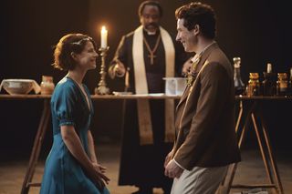 Juliet (Jessie Buckley) and Romeo (Josh O'Connor) smile warmly at each other while the Friar (Lucian Msamati) officiates their wedding.