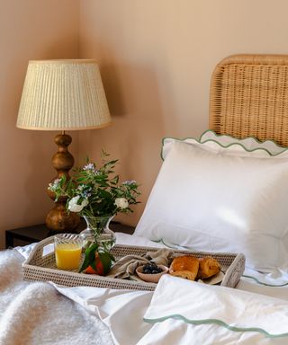 Bedroom detail with rattan breakfast tray on bed, laid with fresh flowers, pastries, juice and berries. Scalloped bedlinen, rattan headboard, and shapely wooden bedside lamp.