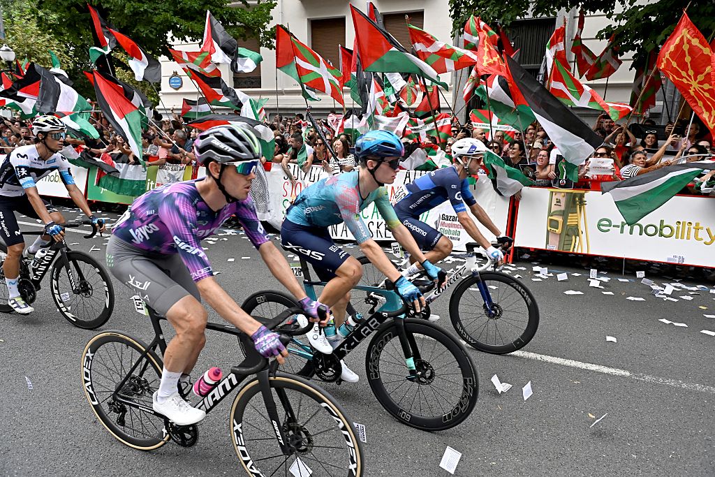 Cyclists compete as pro-Palestinian protesters holding Palestinian and Basque flags demonstrate during the eleventh stage of the Vuelta a Espana cycling tour, a 167 km race from Bilbao to Bilbao, on September 3, 2025. (Photo by ANDER GILLENEA / AFP)