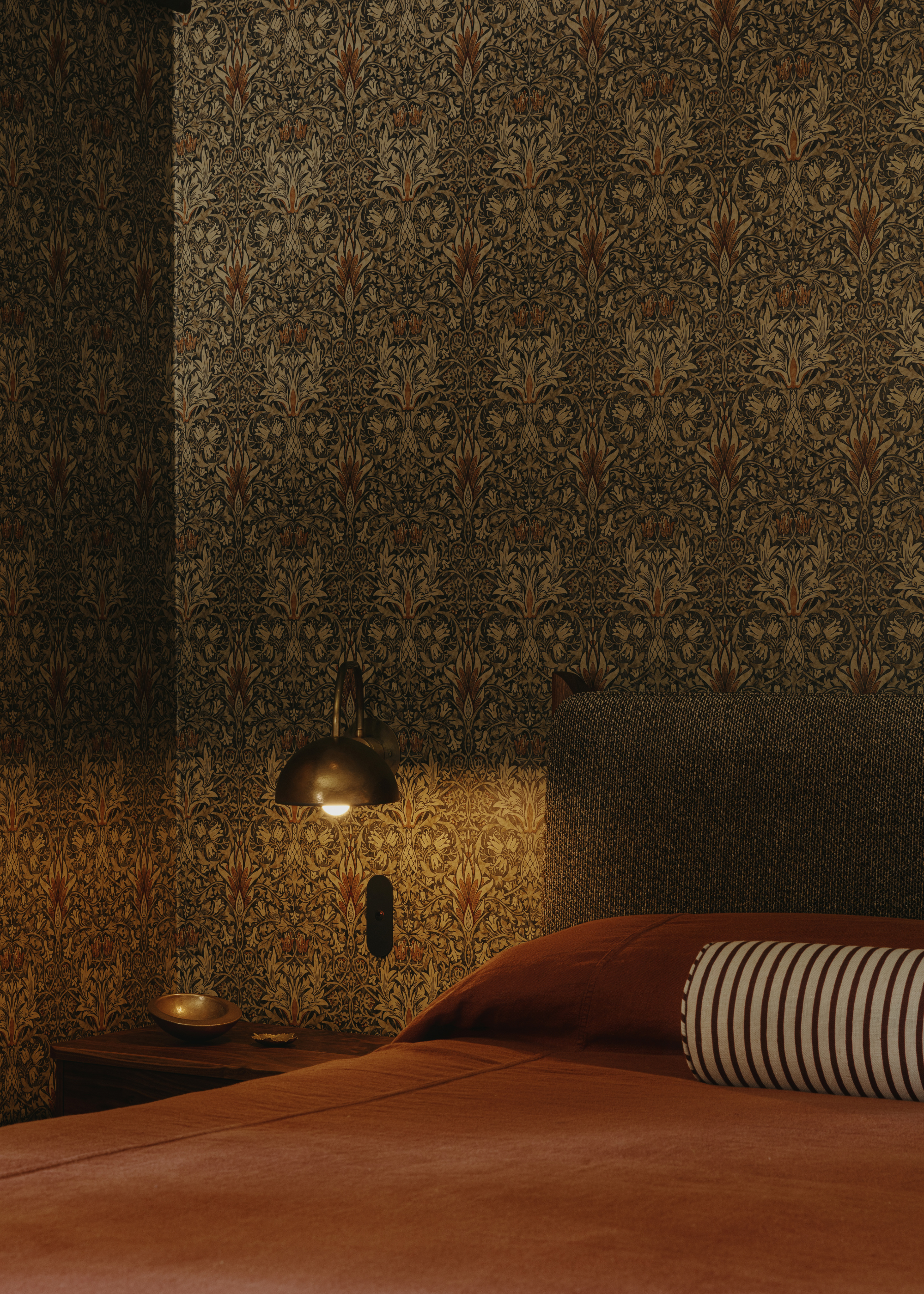A bedroom with patterned wallpaper, a brass wall sconce, a side table with a bowl, a burnt orange duvet, and a striped bolster pillow