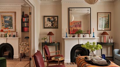 A living room with warm white walls, a fireplace with a mirror above, artwork on the walls, and an orange patterned ottoman and two burgundy armchairs. 