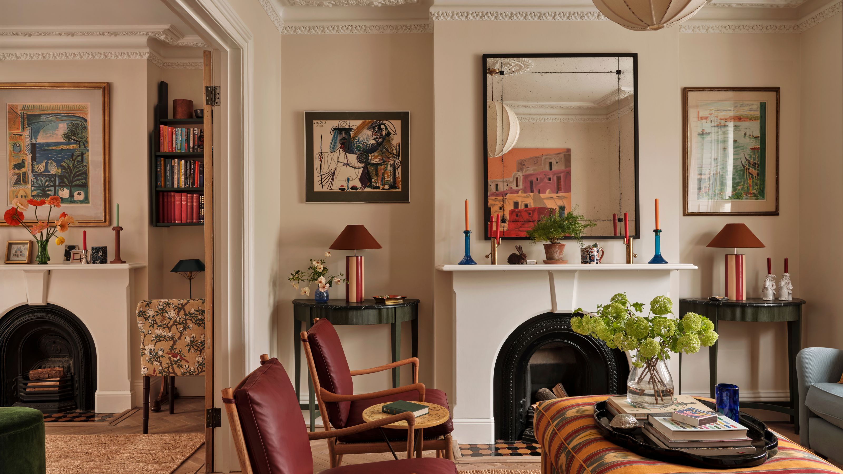 A living room with warm white walls, a fireplace with a mirror above, artwork on the walls, and an orange patterned ottoman and two burgundy armchairs. 