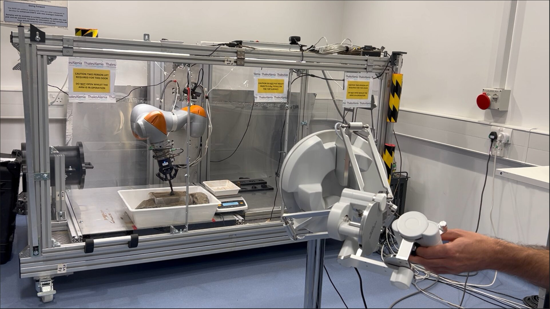 photo of a hand operating, at a distance, a dirt-scooping robot inside a laboratory