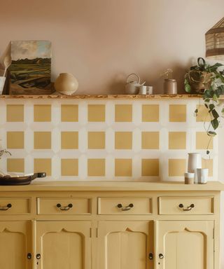 A kitchen with white and yellow grid wall tiles, a wooden shelf above these with plants and wall decor on top, and a yellow cabinet with four drawers and doors