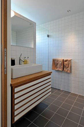 Interior view of the master bathroom at Trekronekabin featuring a white ceiling, white tiled walls, grey tiled floors, a wall-mounted mirror, a white sink on a white and wooden unit and a towel rail with brown towels