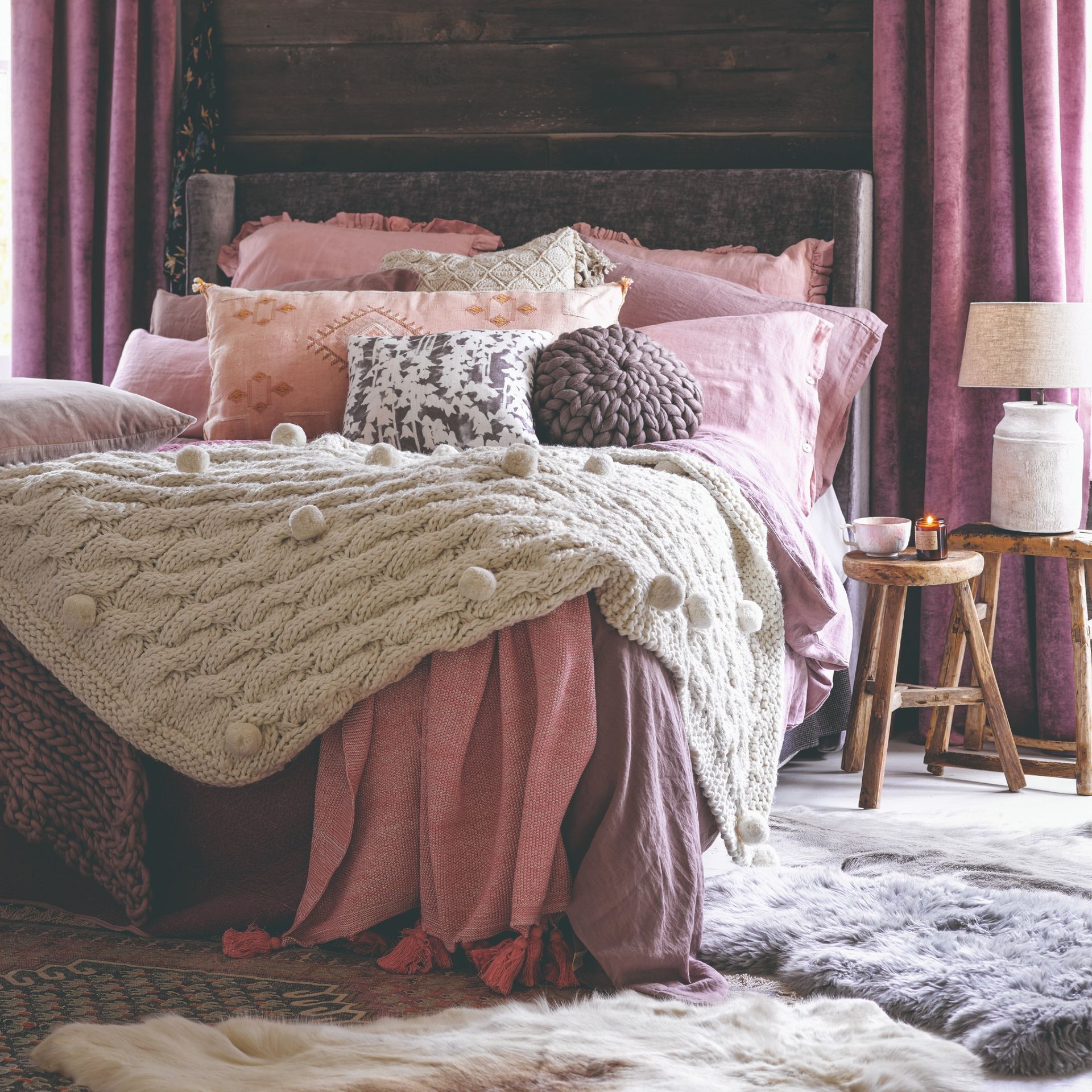 A rustic bedroom with pink bed linen and chunky wool knit throws and cushion with sheepskin rugs on the floor
