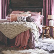 A rustic bedroom with pink bed linen and chunky wool knit throws and cushion with sheepskin rugs on the floor