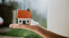 picture of small ceramic house with orange roof balanced on the palm of a hand with sunlight peaking through the window behind to support an article to answer what is Gazundering