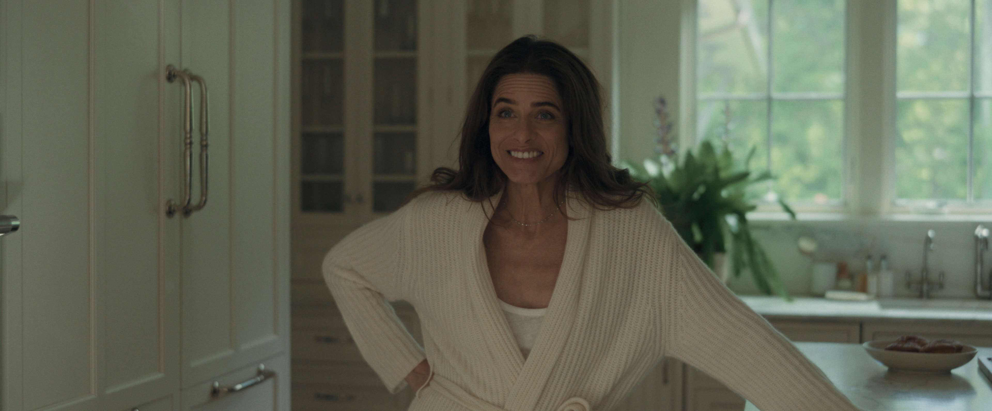 amanda peet as mel cooper smiling an uncomfortable smile and wearing a white robe in a kitchen in your friends and neighbors season 2