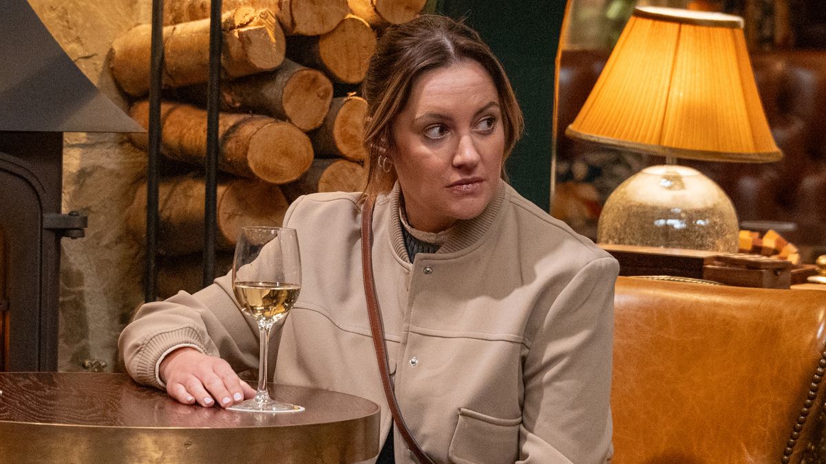 Ella wearing a cream jacket and drinking a glass of white wine in the Woolpack alone and looking nervous 