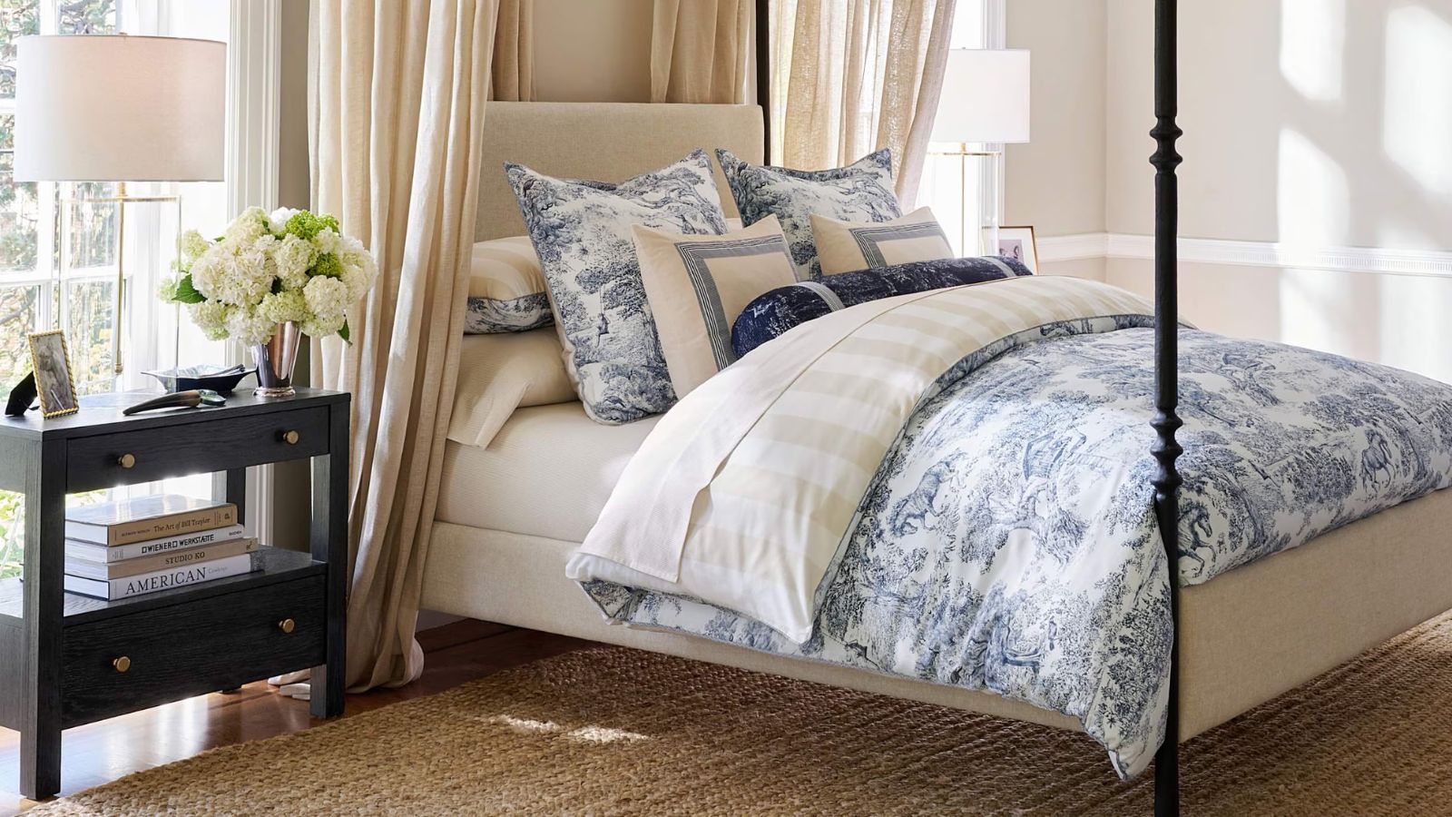 Neutral modern bedroom with four-poster bed, cream bed, blue and white bedding, black side table with lamp, books ans flowers, beige rug and curtains