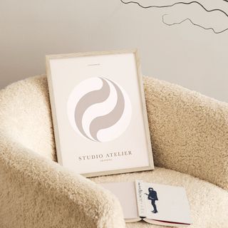 framed picture with a circle logo and studio atelier written on it, sitting on a fluffy white armchair alongside a book