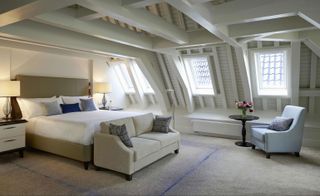 Waldorf Astoria cream colored bedroom with attic roof, exposed beams, cream sofa, light purple armchair and white linen