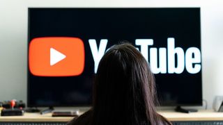 A woman seen sitting in front of a tv screen that shows the logo of You Tube platform