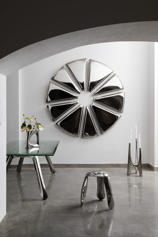 Image of a white room with shiny concrete flooring. There is a dining table with a glass top and legs that look like a deflated chrome balloon. On the table is a chrome vase in the same deflated style. There is a matching stool and candle holder in the room as well. On the wall is a chrome art piece in the shape of a pinwheel.