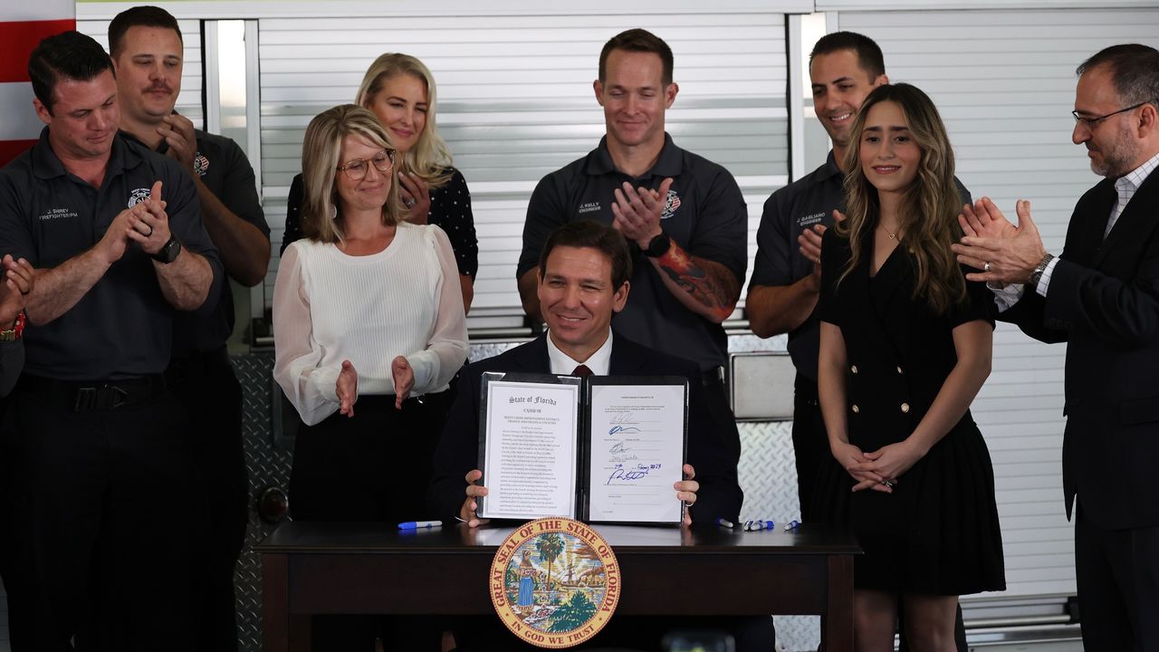 Ron DeSantis behind podium, smiling, holding up signed bill with colleagues smiling and clapping behind him