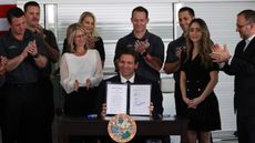 Ron DeSantis behind podium, smiling, holding up signed bill with colleagues smiling and clapping behind him