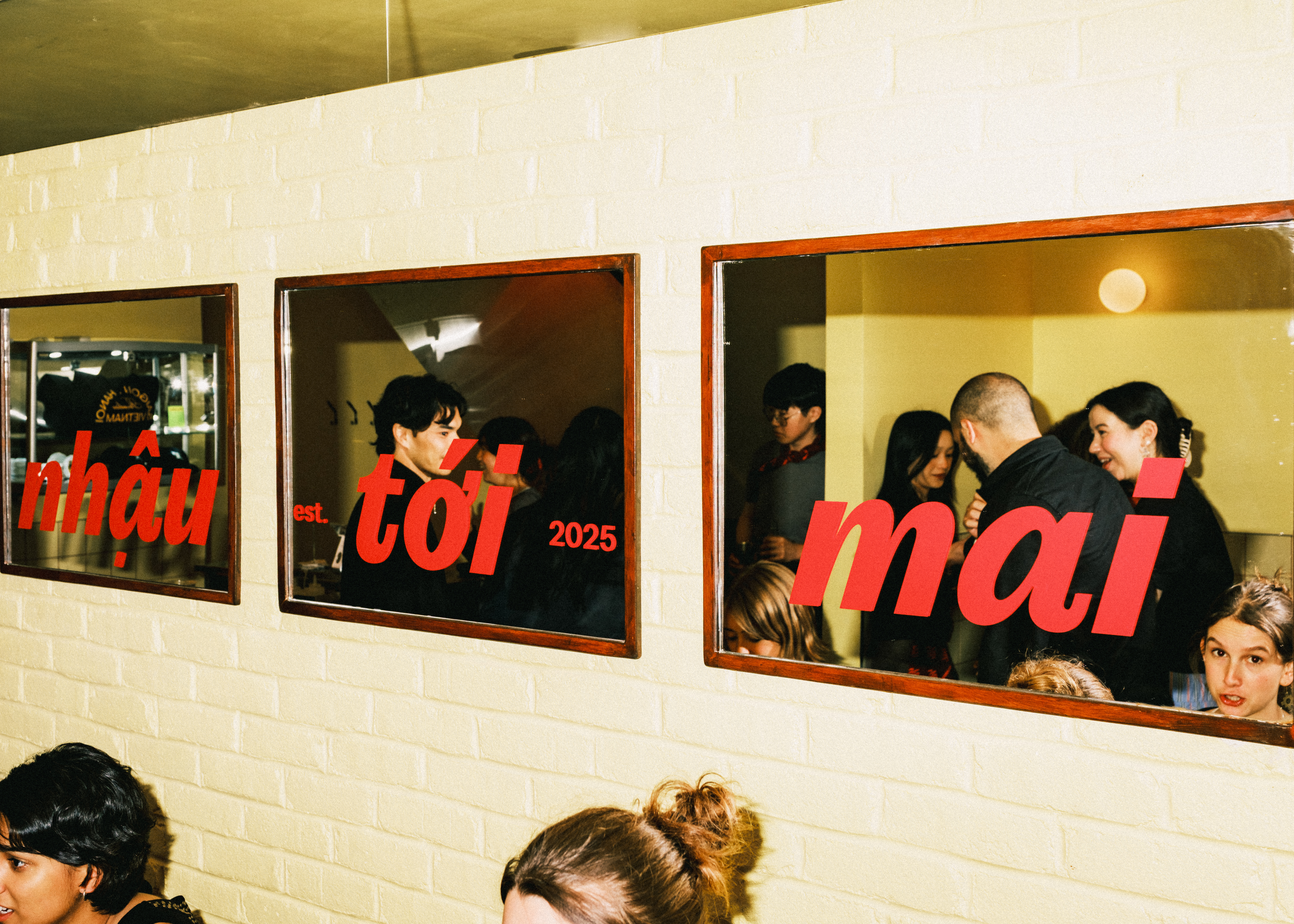 A crowd of people is captured with a film camera mingling inside a restaurant decorated with wooden framed mirrored, brick buttery walls, and red lettering.