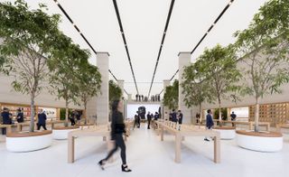 Alternative interior view of Apple's London flagship store featuring light coloured flooring, pillars, long wooden display tables with products on top, trees in round planters and a large screen at the back. There are multiple people inside the store