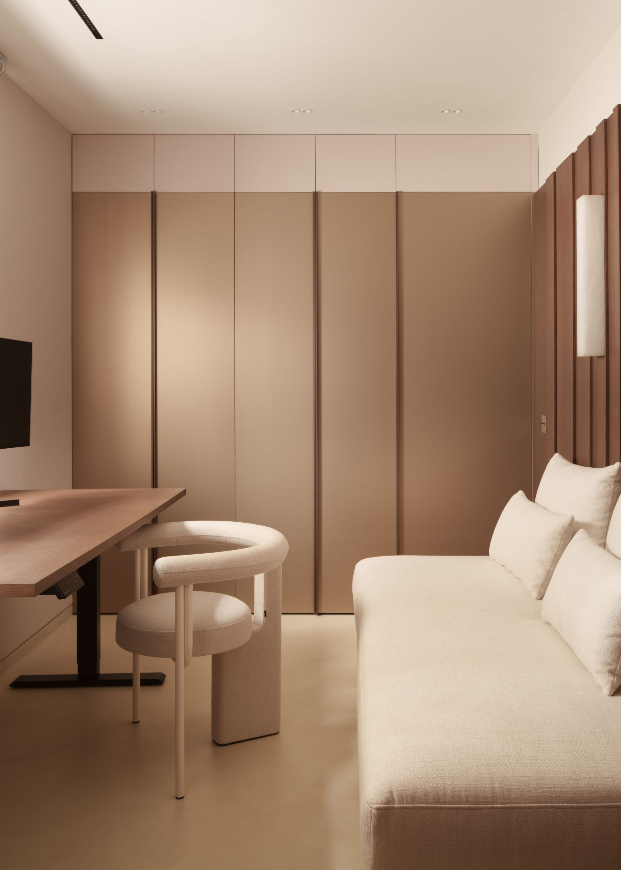 A neutral-hued modern home office with built-in storage on the back wall, a desk against the side wall with a cream curved chair and a cream daybed sits against the opposite wall under a wall light