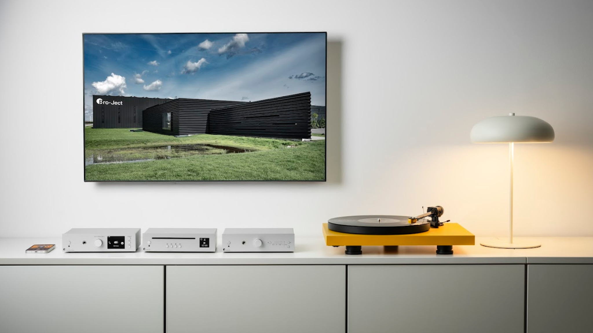 Lifestyle photos showing the Pro-Ject Uni Box S3 on a white unit with a mobile phone playing Massive Attack and a yellow turntable to the right of the amplifier