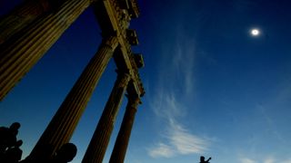a solar eclipse is seen high in the sky above ancient greek ruins