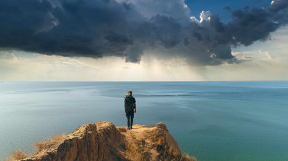 A man stands on a cliff overlooking the ocean and cloudy skies.