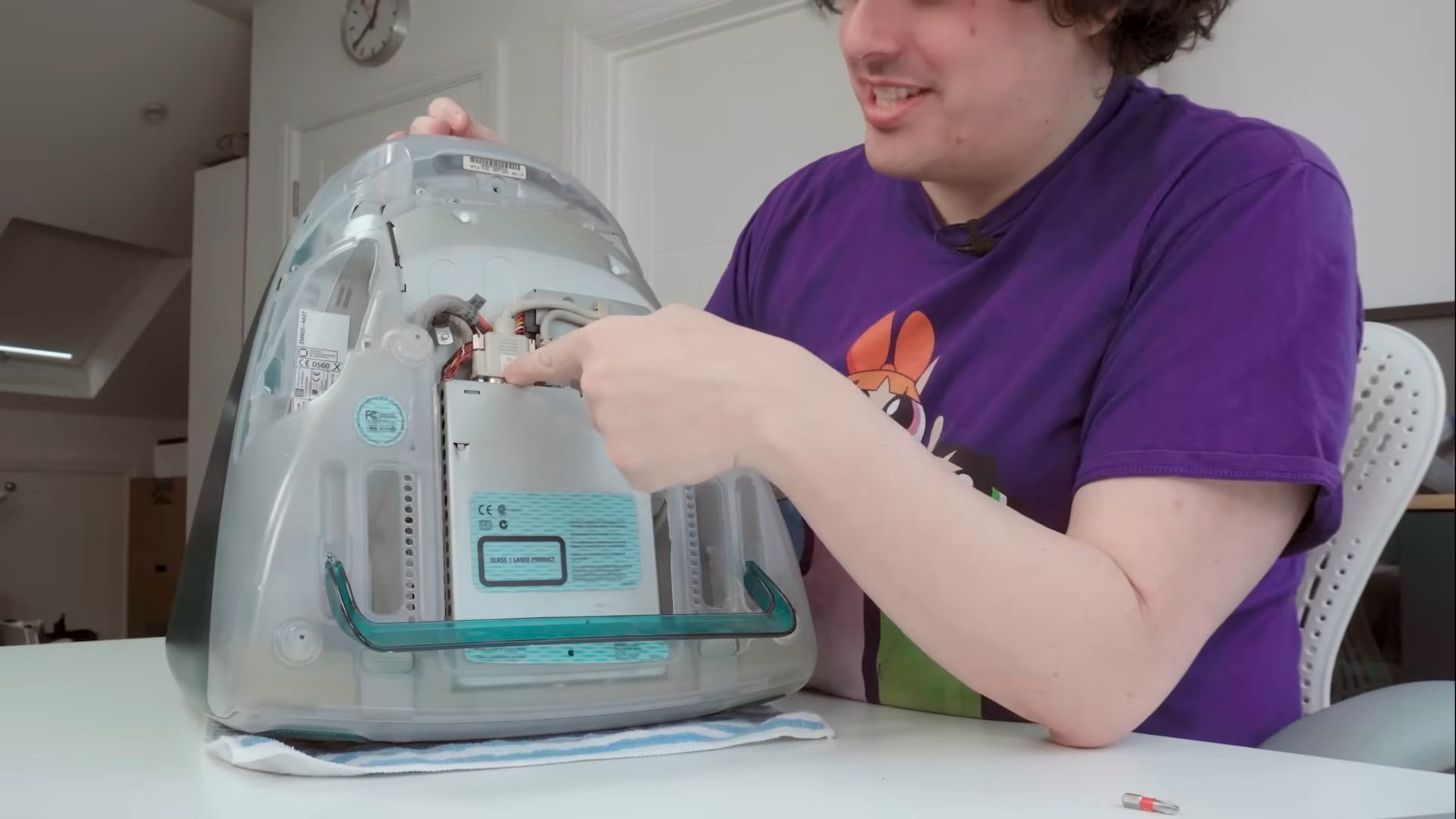 Matt Gray at white desk pointing to iMac G3 video cable with computer on its face and bottom panel removed.
