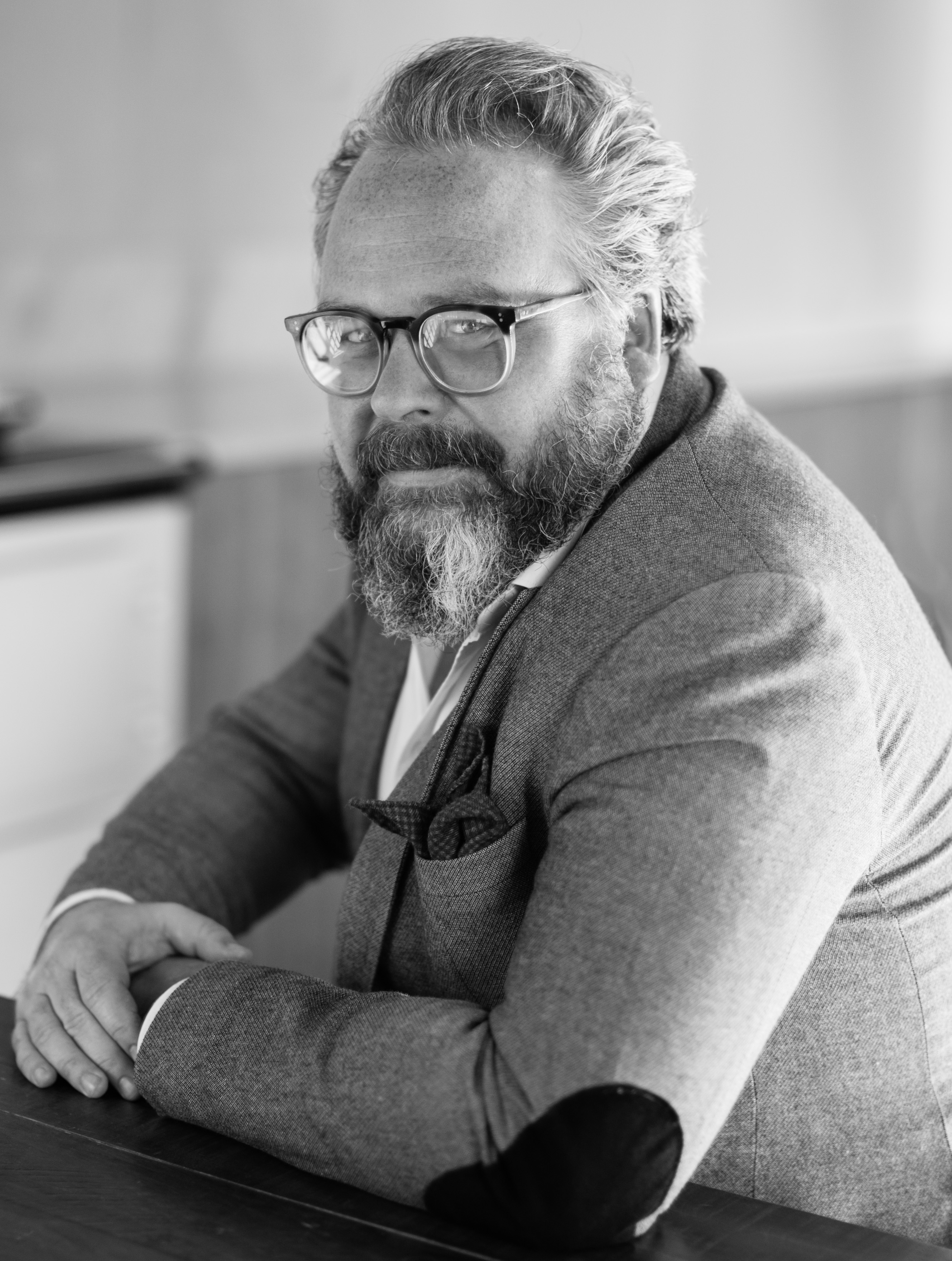 A grayscale headshot of a man with glasses and a beard wearing suit jacket 