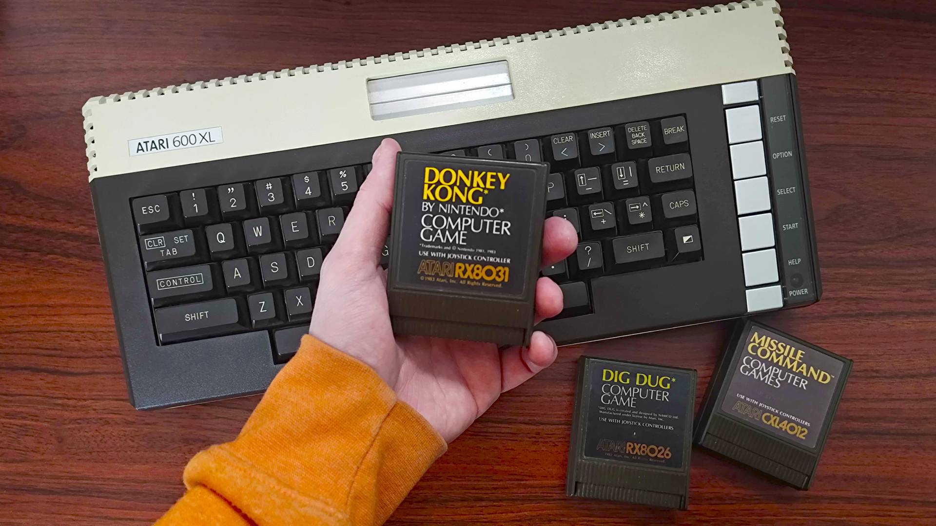 Hand holding Donkey Kong cartridge in fromt of Atari 600XL 8-bit micrcomputer on woodgrain desk.