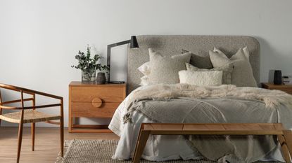 A grey fabric bed with lots of throw cushions, with an oak bedside table 