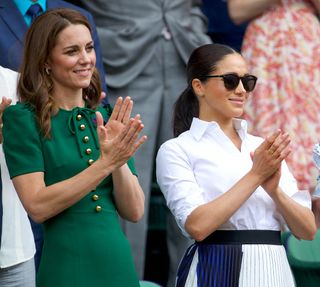 Catherine, Duchess of Cambridge stands with Meghan, Duchess of Sussex in the Royal Box on Centre Court