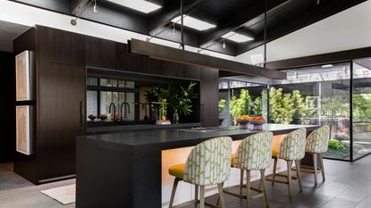 Example of kitchen ceiling ideas. Large kitchen with black wooden ceiling and skylights, large black waterfall kitchen island with white base, four upholstered white and green bar chairs, large metal task light above island, dark wooden cabinets, dark gray flooring, large floor to ceiling windows looking out to garden space.