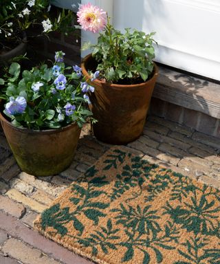 A dark brown brick wooden porxh with a brown doormat with green leaf illustrations and two potted plants in brown pots next to it with blue and pink flowers, in front of a white door